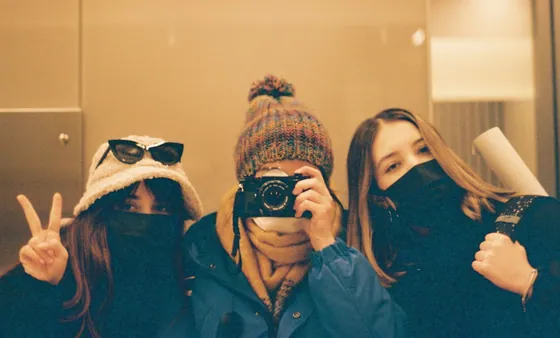 Three friends in winter coats and knitted hats crowded together for a photo, the one in the middle holding a film camera up to her face and the one on the left throwing a peace sign